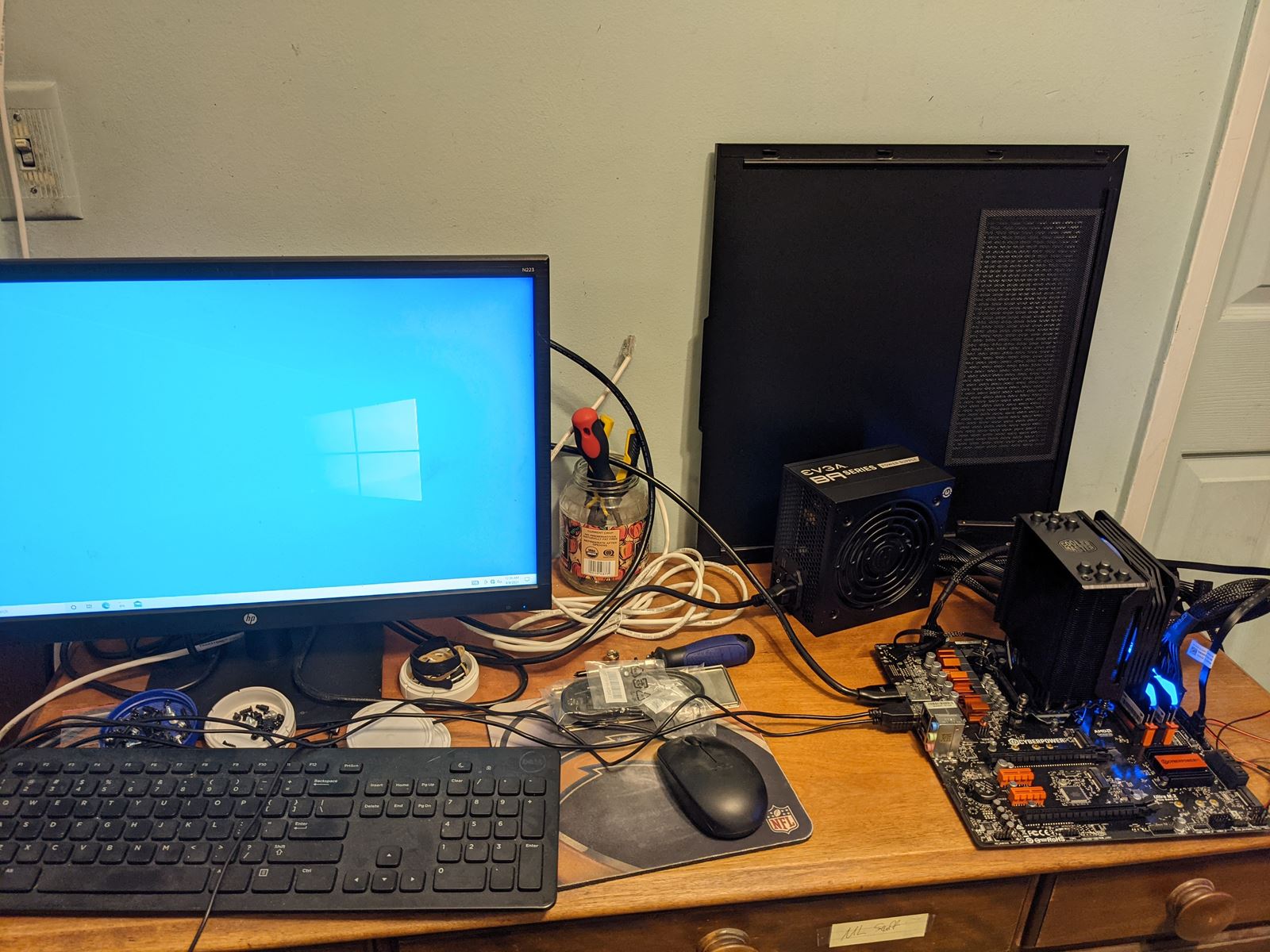 A workbench with a monitor showing the Windows desktop, a keyboard, a power supply and a motherboard with a tower cooler being tested outside its case.